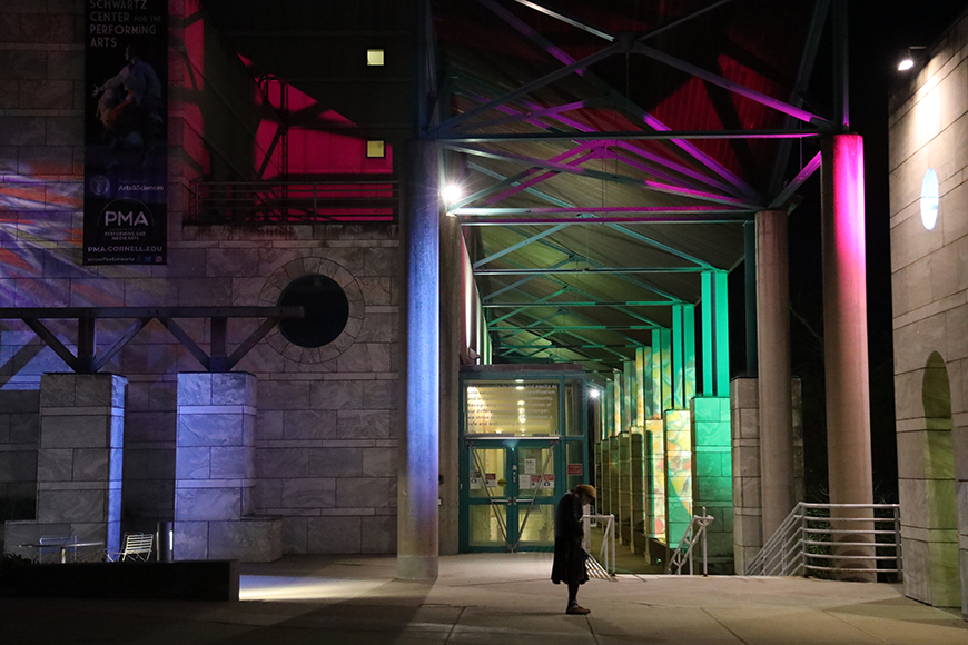 A view of a large, grey brick building at night. Red, purple, pink, and green light projects onto the front of the building's facade, consisting of grey brick and green metal support beams.
