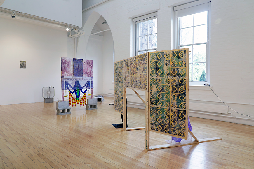 A gallery view of several pieces of art: a small painting on the far left wall; a wooden frame draped in purple and blue fabric, held up on concrete blocks, with colorful balls of blue, red and yellow balls hanging from it; and a wooden screen with a design of green and black squares printed on it.
