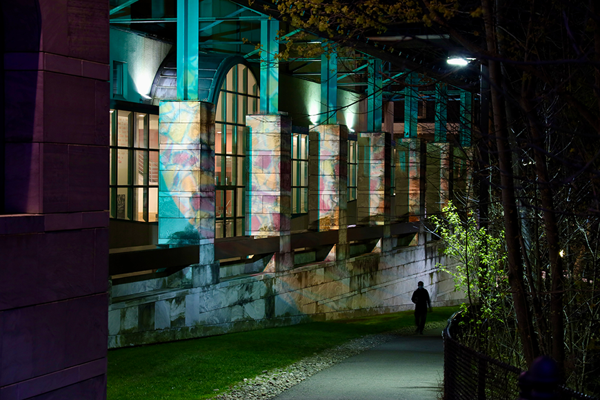 At night, multicolored light, reminiscent of stained glass, projects onto the side of a building's facade, consisting of grey brick and green metal support beams.