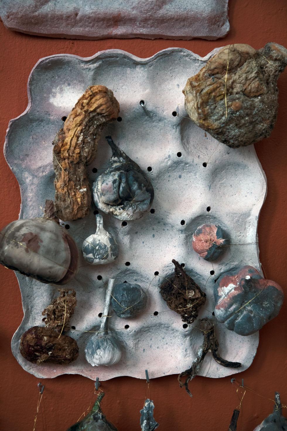 Gray colored egg carton with various clay fired gourds placed throughout set on a dark red wall.