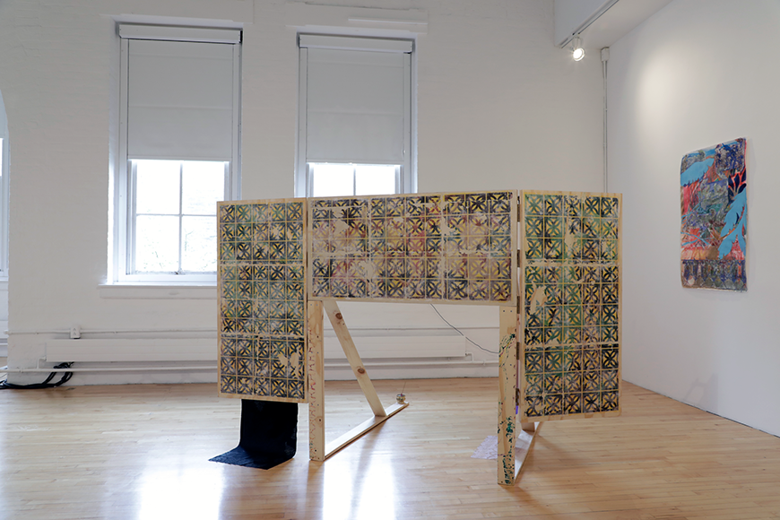 A gallery view of a wooden screen with patterned green and black squares printed on it, and a drawing with blue leaves hanging on the wall in the background.