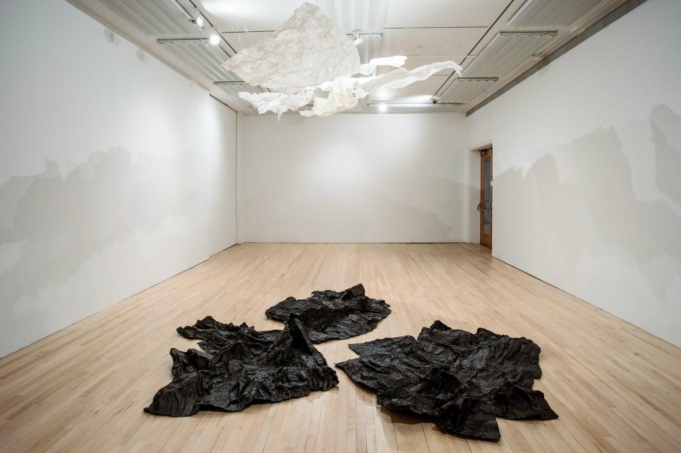 Three large black crinkled papers on a light wooden floor with a large crinkled paper sculpture hanging from white ceiling surrounded by three white walls.