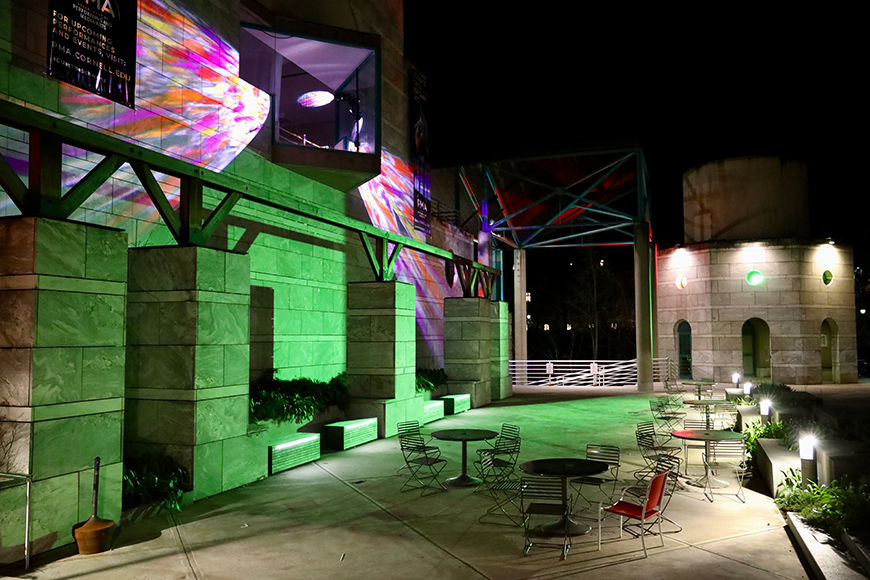 At night, multicolored light, reminiscent of stained glass, projects onto the front of a building's facade, consisting of grey bricks and a large angular bay window on the second story. The projections shine out of the angular window; a few tables and chairs sit outside the building, underneath the projections on a wide sidewalk. 