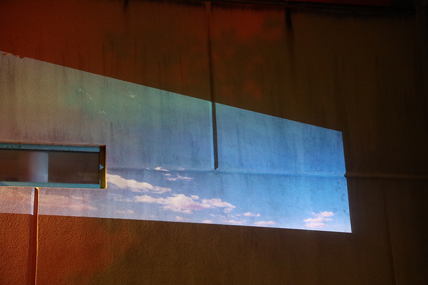 One distorted rectangular projection shines on a grey brick wall and a small rectangular window. The projection is a light blue sky with scattered, wispy white clouds. 