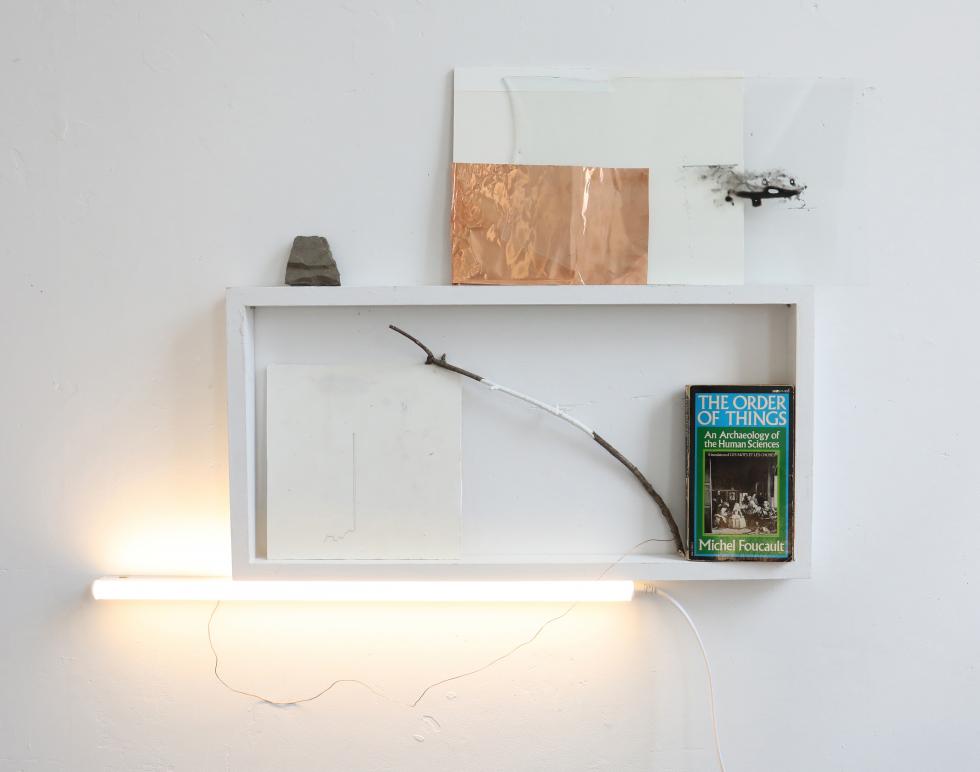 White open box on a white wall with a book, stick, copper foil stone, LED light arranged within, on top of, and beneath it.