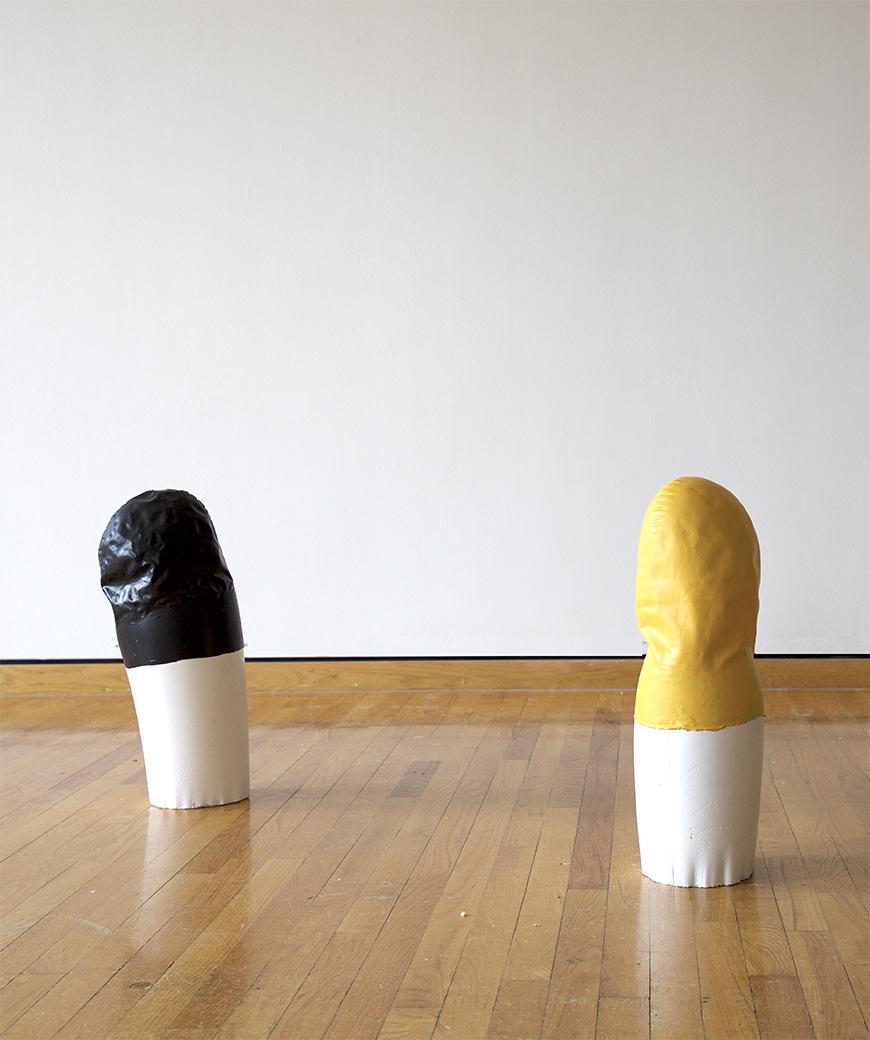 Two abstract sculpture objects, one with a black top half, and one with a yellow top half, both with white bottoms, set on a wooden floor against white wall.