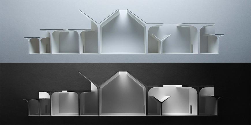 Two photographs of light-study models made out of white chipboard and paper materials showing section-cuts through proposal, with the bottom model being on a darker background and only being lit in the interior spaces. 