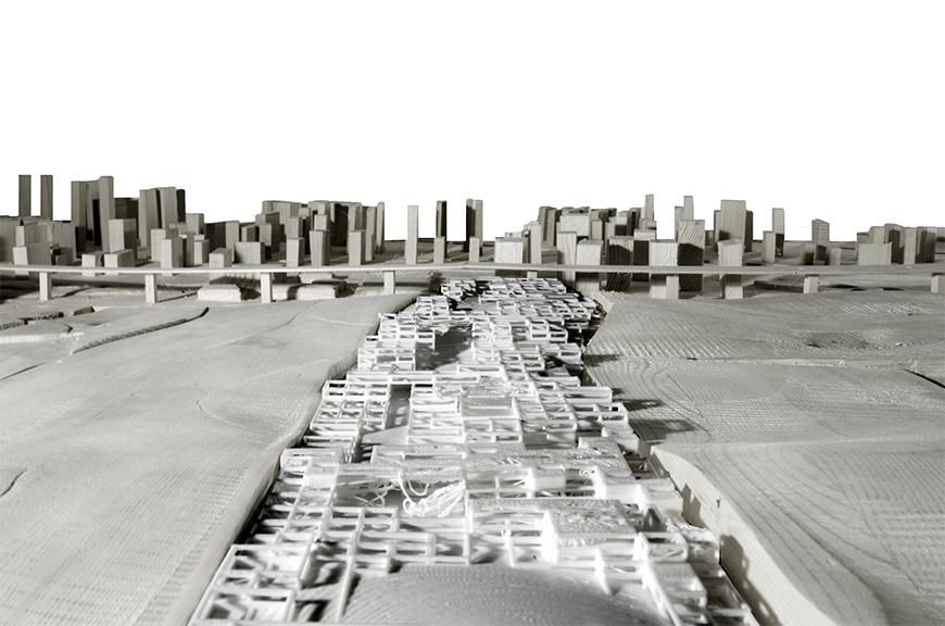 Photograph of white 3D printed model from above image placed into larger wooden site model, shot in a high-angle point of view looking down along the main axis of the model at a skyline in the background.