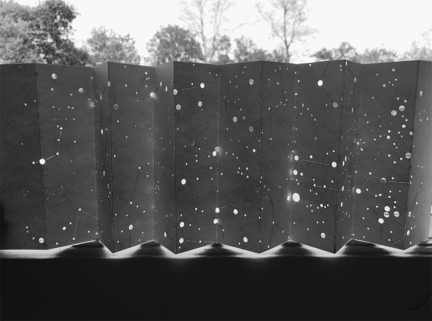 Black and white photograph of map made on pleated paper surface by creating holes to indicate the placement of stars, placed against a window so that light comes in through the openings.