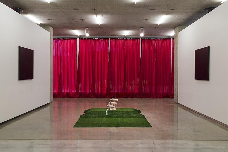 Two white walls with identical dark canvases facing each other, red curtains in the background and a tan abstract sculpture piece on green turf.