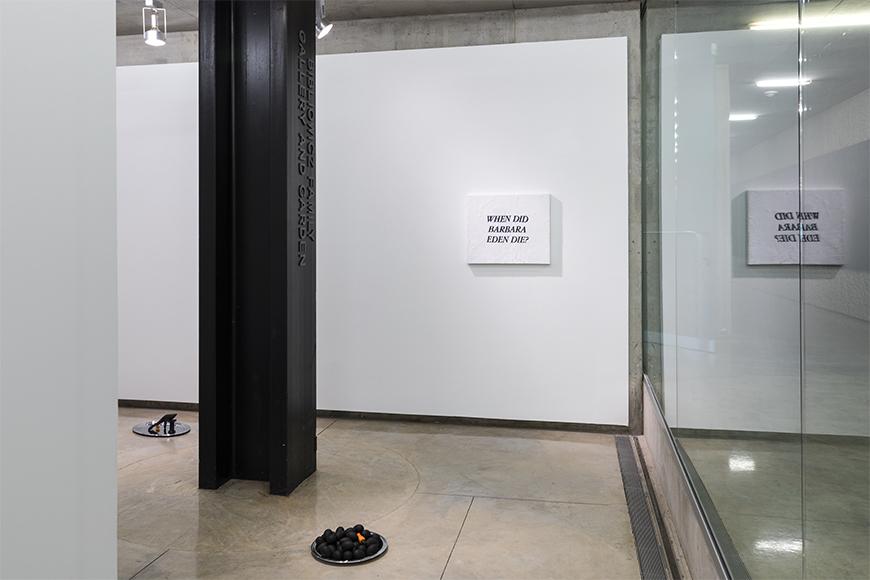 A black pillar in front of a white wall, holding a white sign with black images, mirrored in glass to the right, with a small platter of black objects on the ground.