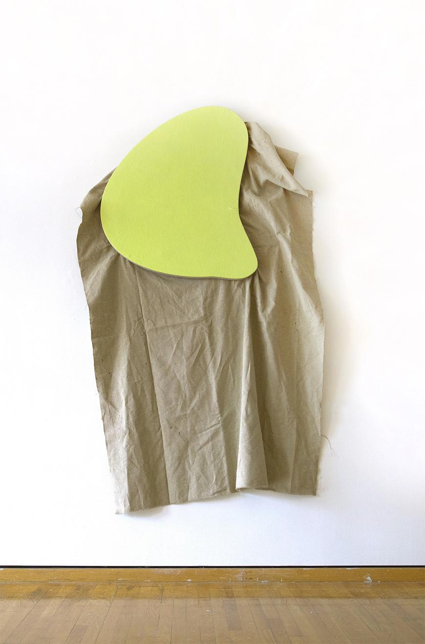 Tan colored fabric hung from a white wall with a yellow green abstract shape holding it up.