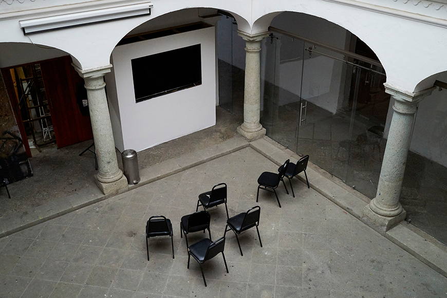 Horizontal color photo of seven black chairs sitting in a concrete and stone atrium. Image taken from an aerial perspective, facing the backs of the chairs. 