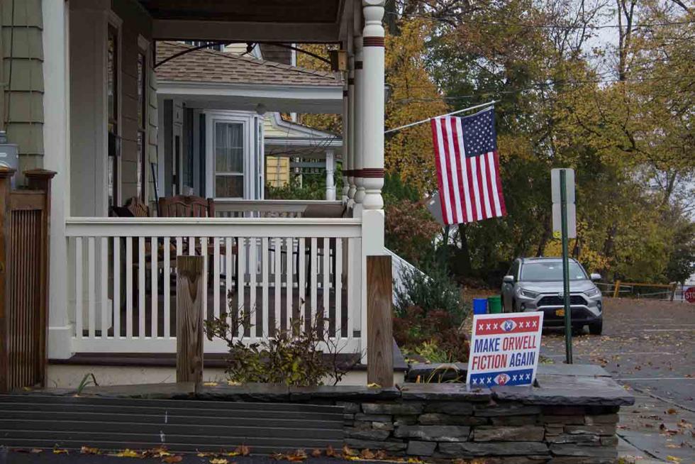 Lawn sign in red, white, and blue, with the text MAKE ORWELL FICTION AGAIN next to a porch with an American flag and parking spots.