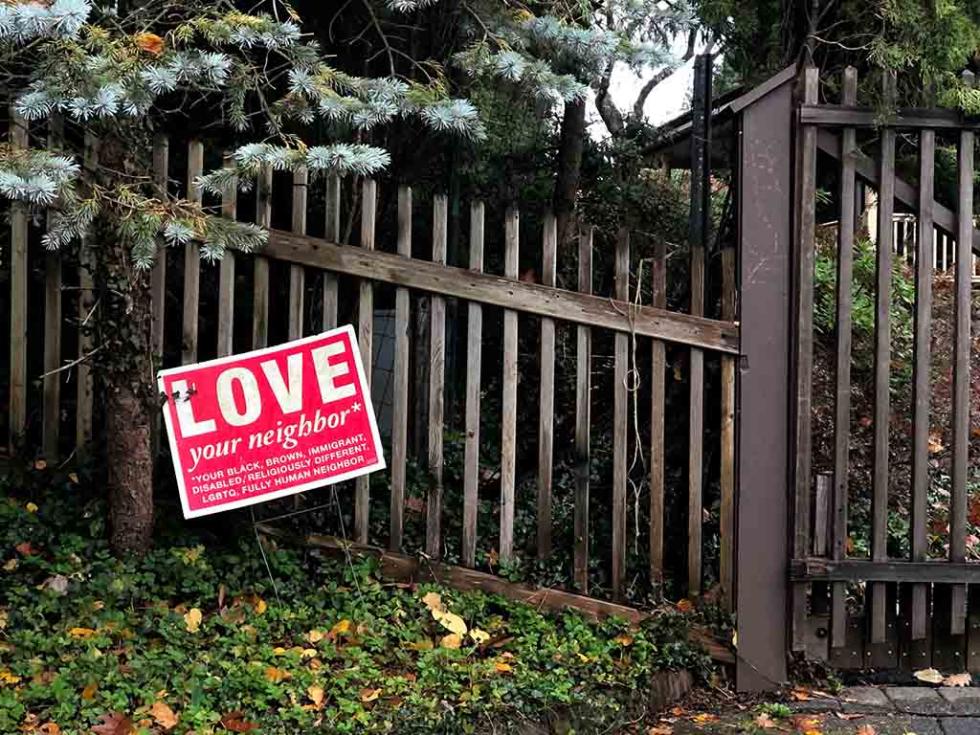 Red and white sign with the words LOVE your neighbor, in front of a brown picket fence on grass.