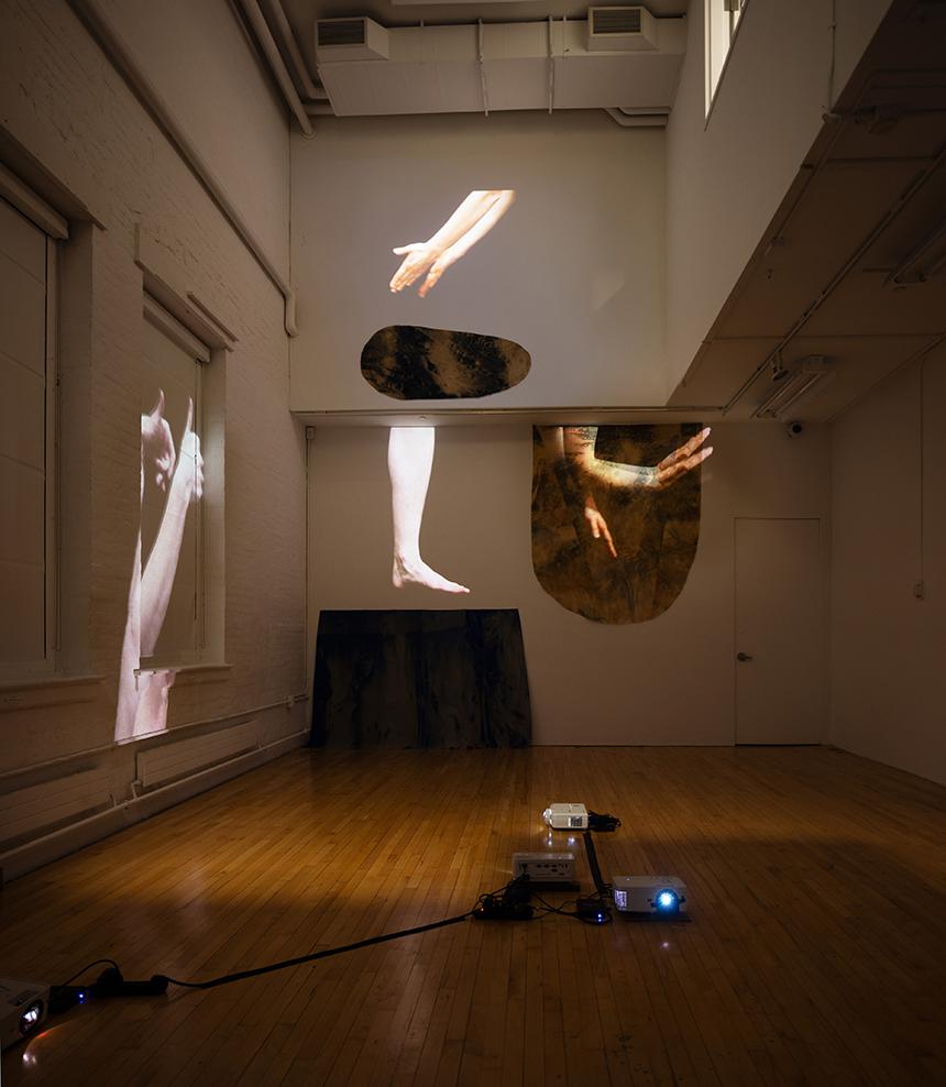 Projected images on a gallery wall of arms and legs posed in interesting positions. The video projector can be seen in the middle of the wooden floor.