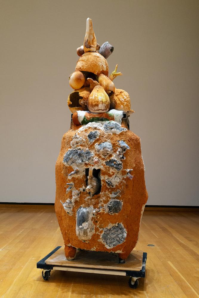 An abstract sculpture sits atop a square transport dolly in a gallery with light wood floors and white walls. The sculpture is mustard, grey, and white, with bulbous, vegetable-shaped forms sticking off of it in all directions. 