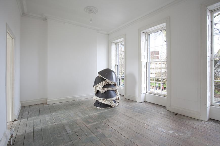 Installation of metallic black domed shapes with tan fabric stacked on top of one another inside a room with bare white walls, natural light coming in through large windows, and grey wooden floors. 