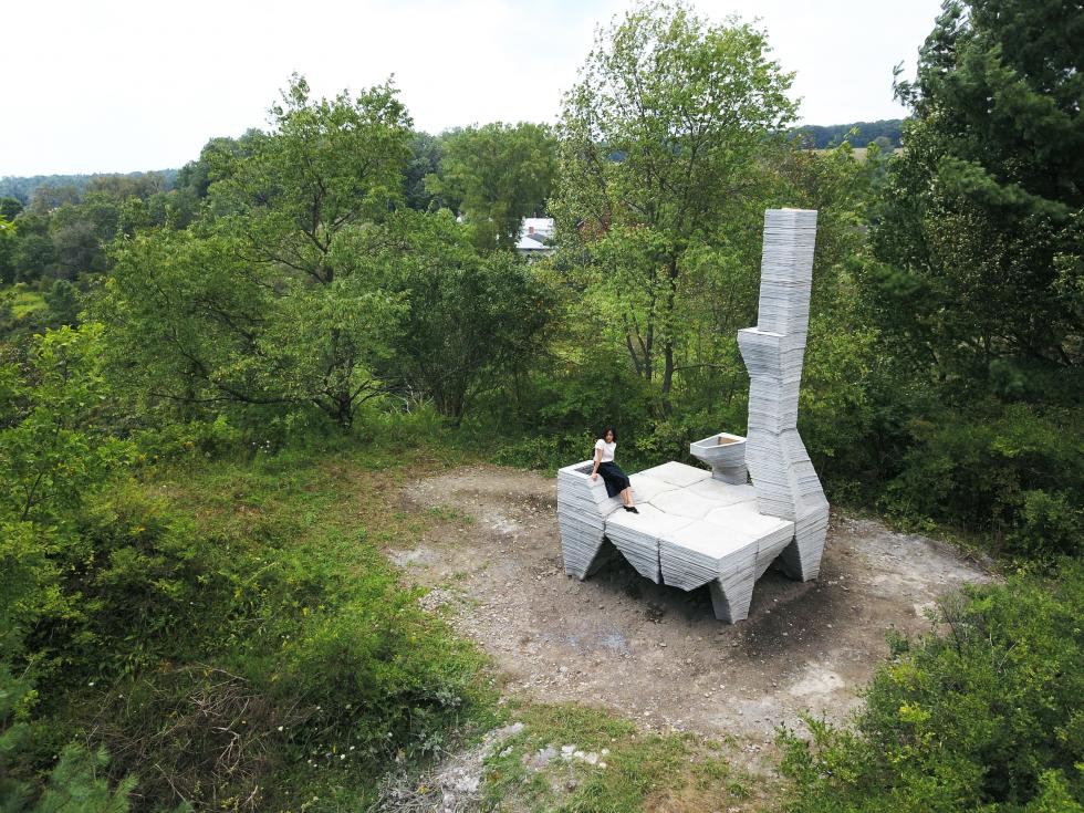 a white structure sitting among trees, with a woman wearing black and white with dark hair sitting on top of the structure