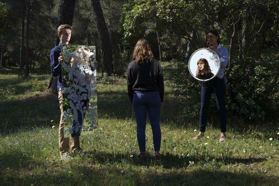 Three people outside in greenery with two people holding up floral artwork and a mirror with one person in front staring at them.