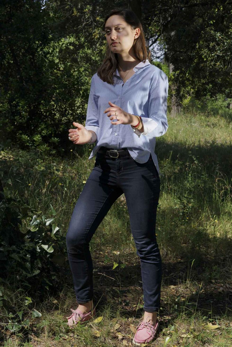 Woman wearing a light blue blouse with dark jeans outside in nature with their hands outstretched.