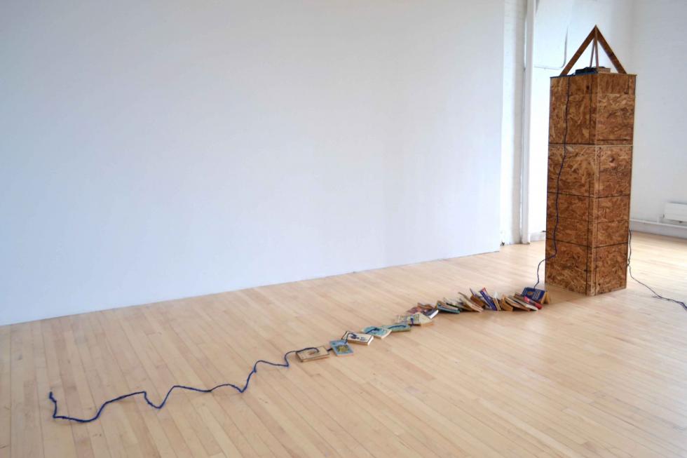 Single obelisk with blue string connecting a series of books together on the floor in a white room.