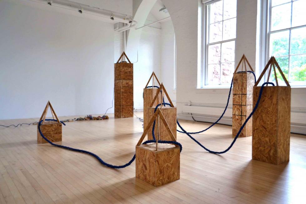 Wooden obelisk statues of various size with blue rope connecting them together in a white walled gallery with a light wood floor and long windows which bring sunlight in. 