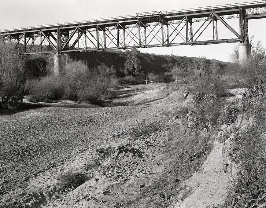 A black and white landscape of a dry river bed with shrubs interspersed throughout and a wooden cross in the distance. Above, an elevated bridge stretches out of sight in both directions.