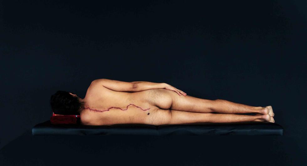 Man in the nude laying sideways with a jagged red line going from his neck to tailbone representing US Mexican border outline.
