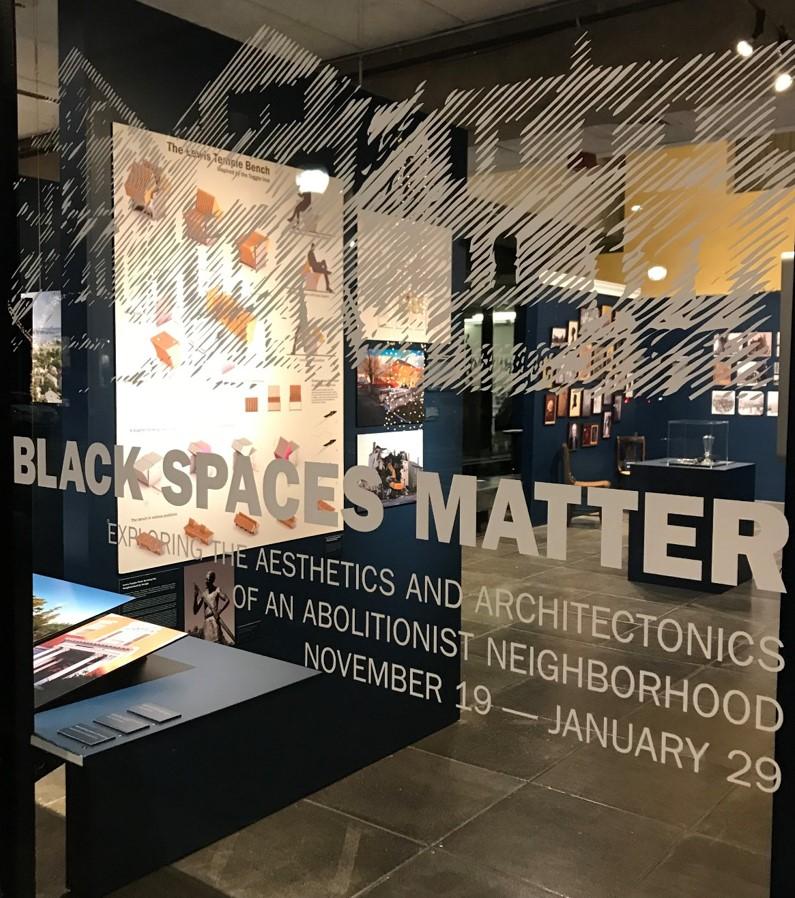 Exhibition space with a focus on 'Black Spaces Matter.' The glass window has white text displaying the exhibition title, 'BLACK SPACES MATTER,' and details, 'Exploring the Aesthetics and Architectonics of an Abolitionist Neighborhood, November 19 — January 29.' Inside, various displays and informational boards are visible, showcasing images, diagrams, and artifacts related to the theme.