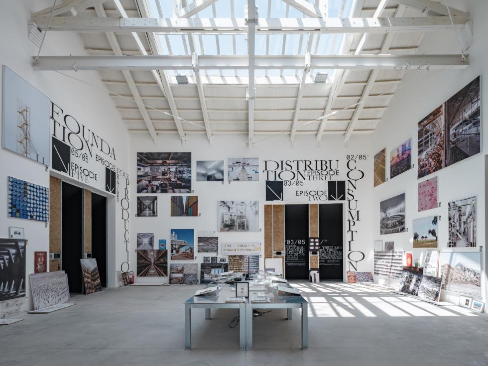 A large, open gallery space with a white vaulted ceiling and skylights. The walls are covered with framed posters and images of architectural plans and models. Tables display additional materials.