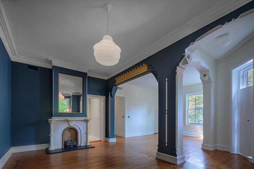 A photo of a spacious, historic living room with a white marble fireplace, dark blue walls, and ornate white trim. There is a large white pendant light hanging from the ceiling and a mirror reflecting the room on the left wall. A doorway leads to another room on the right, and a large window with white curtains is visible in the background.