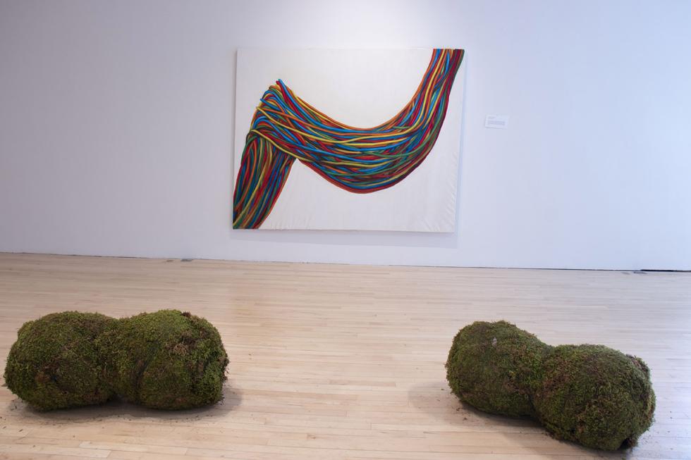 Rainbow colored painting of multi-colored strings draped across the canvas behind two moss covered boulders.