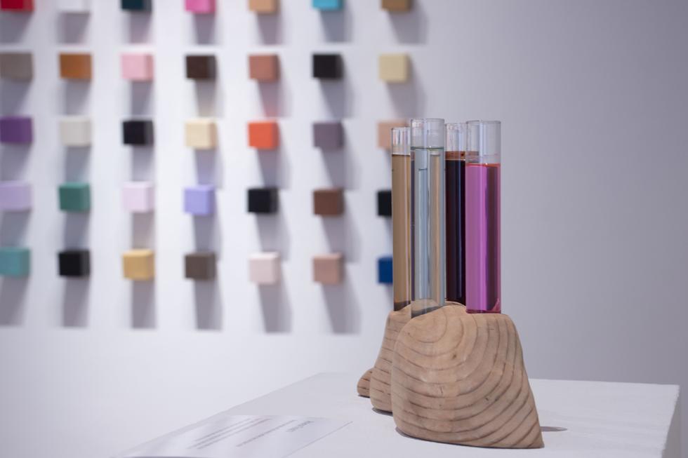 Multi-colored vials of liquid in a small wooden holder with rings on it in front of small colored squares hung on the wall.