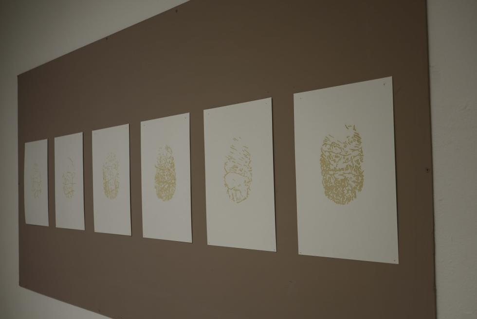 Six prints of tan ink on white paper are posted side-by-side on a light bulletin board. The six abstract prints are all different from each other but share the same dimensions. 