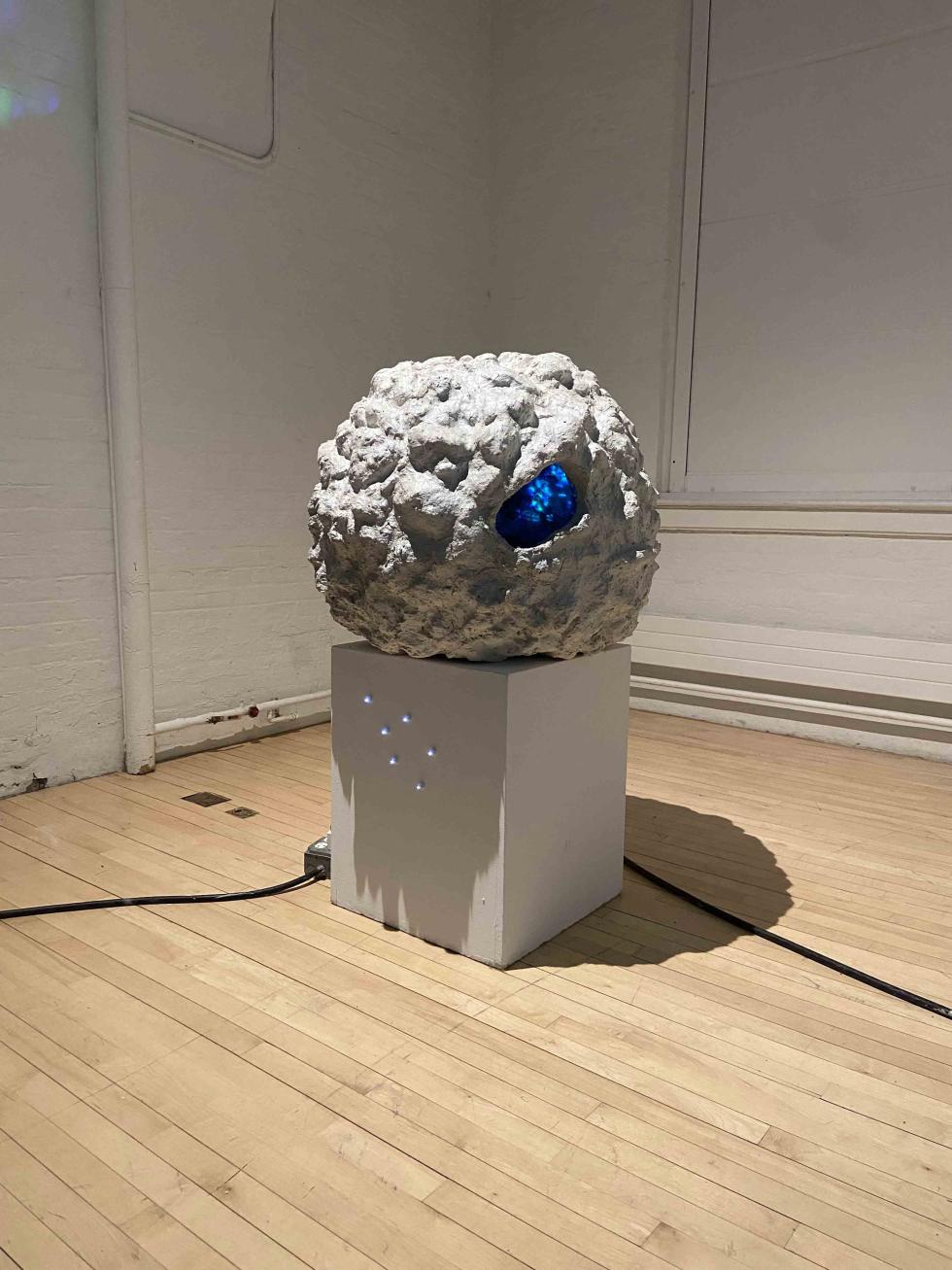 Inside a gallery with white walls and a light wood floor sits a white pedestal. Atop the pedestal is a sculpture reminiscent of the moon; grey and rocky, with a hole in it the exudes a blue light.