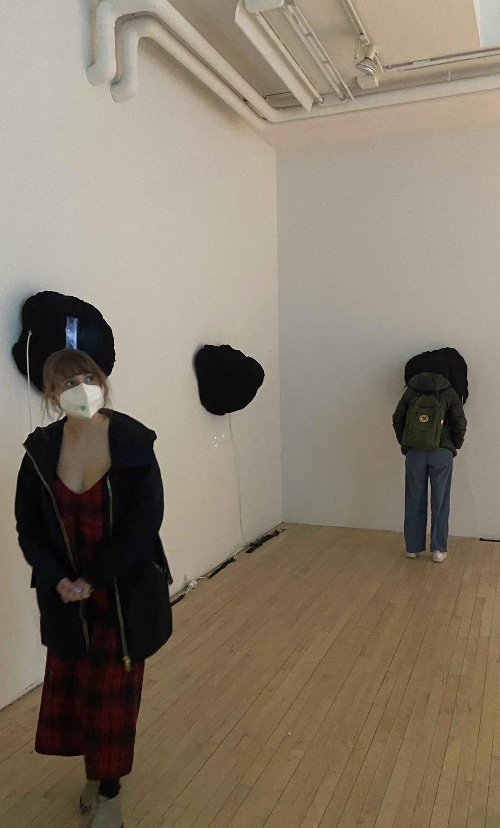 Inside a gallery with white walls and light wood floors, a person stands with their back to the camera and looks closely at a black mass sculpture affixed to the wall. Facing the camera and looking across the gallery is a person wearing a red dress, black sweater, and white face mask. 