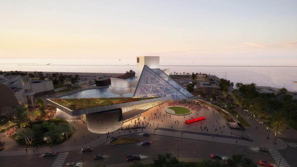 Aerial view of the Rock & Roll Hall of Fame at sunset. The iconic pyramid-shaped building is illuminated, with visitors walking around the plaza and enjoying the outdoor spaces. The waterfront and cityscape are visible in the background.