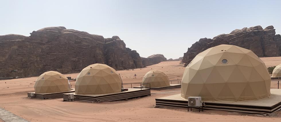 A cluster of geodesic dome tents set up on wooden platforms in a desert landscape. The domes are beige with small circular windows and are equipped with air conditioning units. The surrounding area features vast sandy terrain and towering rocky cliffs under a clear sky.