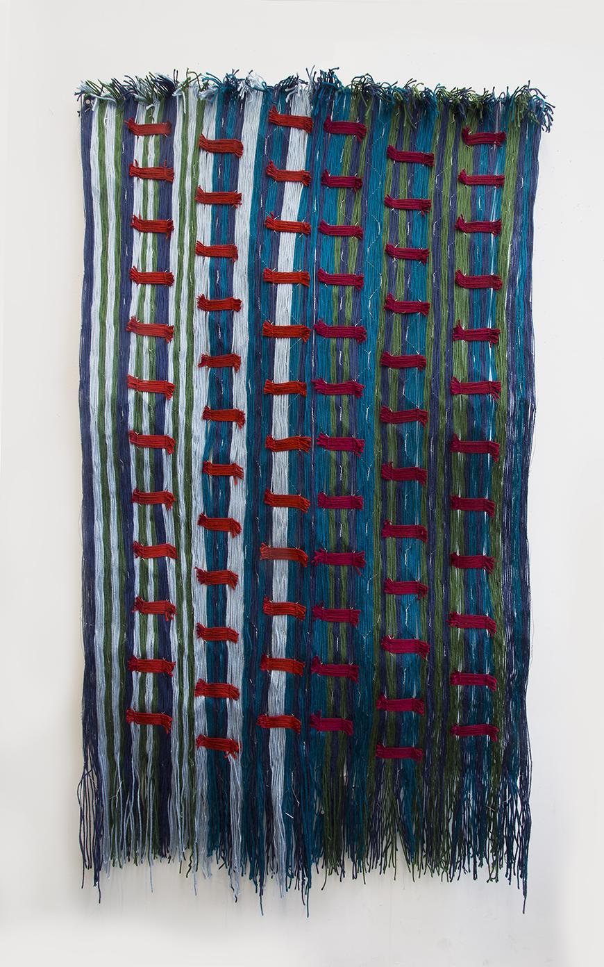 Photo of a fiber sculpture: Strands of light blue, navy blue, teal, and olive green yarn are woven together in a vertical stripe pattern, with irregular stripe widths. Many segmented maroon colored horizontal stripes interrupt the vertical stripe pattern in regular distances. 