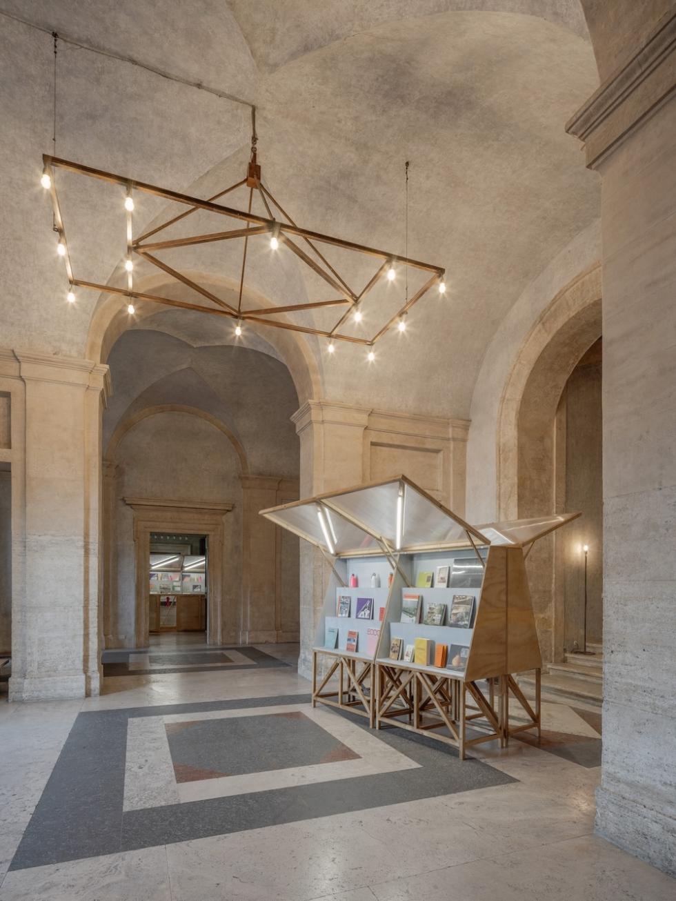 A modern, wooden book display stands in a spacious, arched hallway with marble floors. A large, geometric light fixture hangs from the ceiling above.