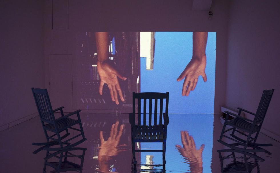 Empty room with three rocking chairs in front of a projection of two arms reaching down in front of a sky and building background.