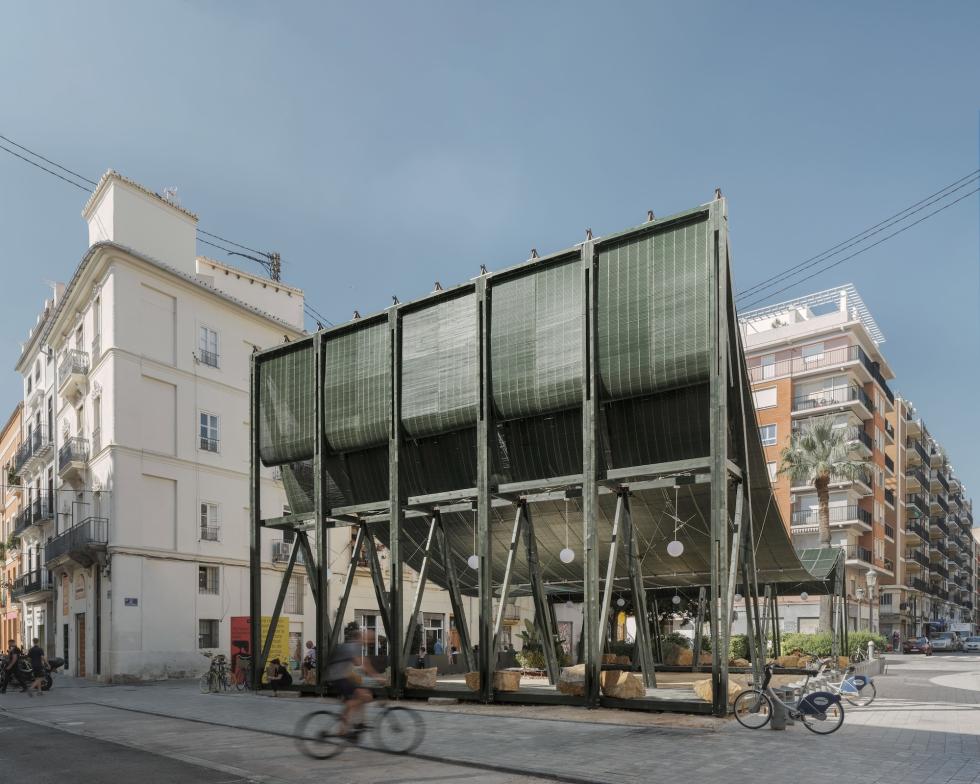 A modern, open-air pavilion with a green metal roof stands in a city square. The pavilion has a lattice-like structure with wooden benches and seating areas inside. People are walking and cycling on the surrounding streets.