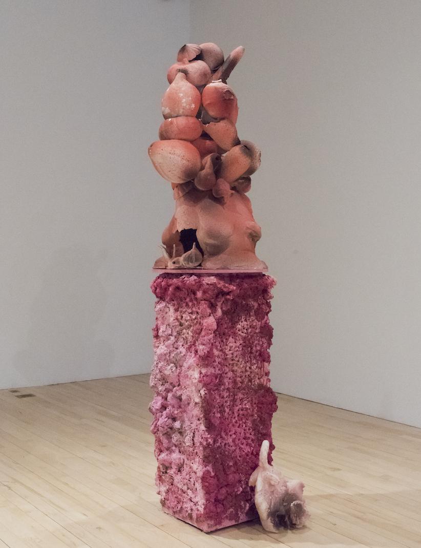 An abstract, rectangular, pink sculpture sits in a gallery with a wooden floor and white walls. The bottom half is a pale pink and fucshia and the top half is peachy pink and light brown, 