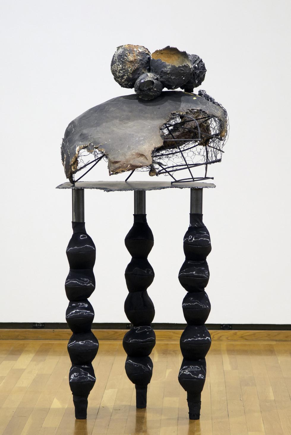 An abstract sculpture with three legs, similar to a small table, sits in a gallery with wooden floors and white walls. The sculpture contains chicken wire, shiny black paint, and rounded forms made of paper maché. 