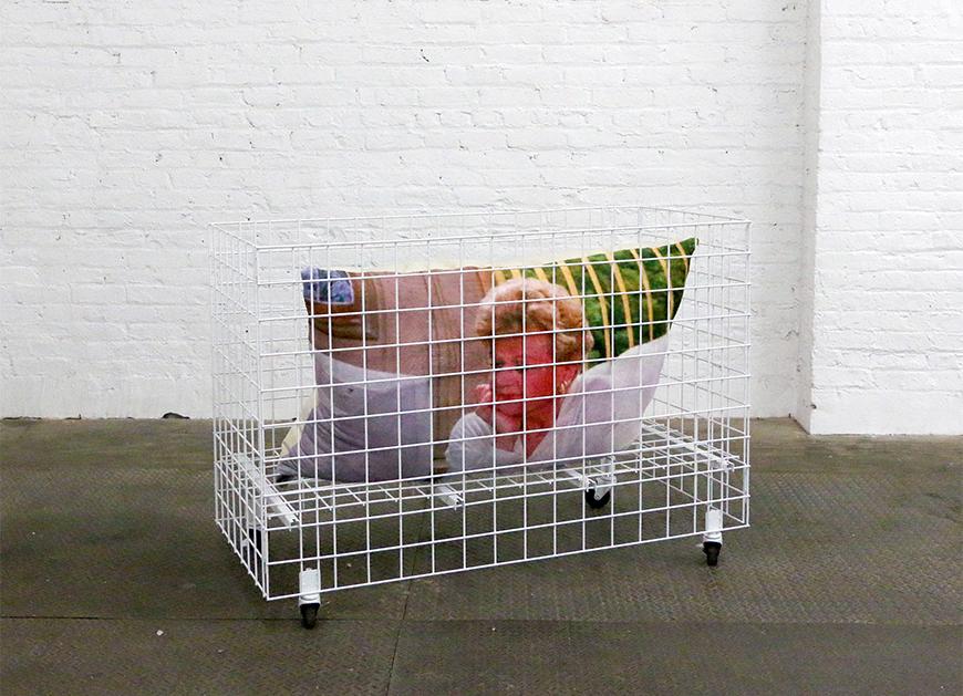 A pillow featuring Angela Lansbury is in a white cage with rolling wheels on the bottom on a concrete floor with a white brick background.