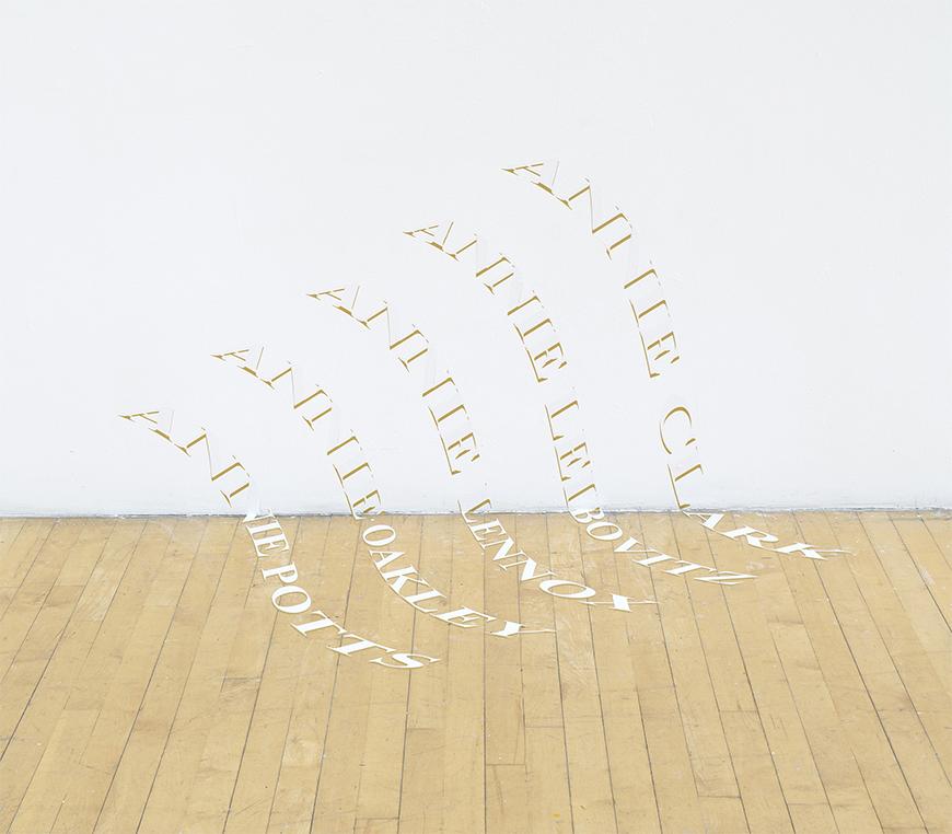 White and gold lettering featuring five different Annie titles waved parallel towards each other going from the white wall to tan wooden floor.