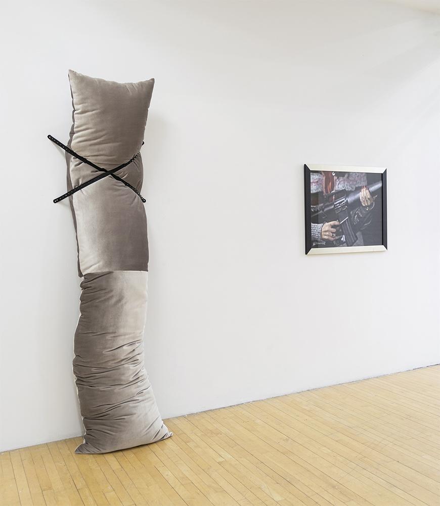Grey velour body pillow propped vertically against a white wall with two black straps securing in an X shape with a black and white framed photo of someone carrying a weapon.