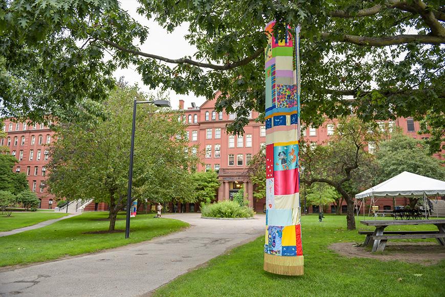 A multi-colored fabric tube that hangs from the top of a tree branch all the way down to the grass. In the background is a large brick building and a path leading up to it.