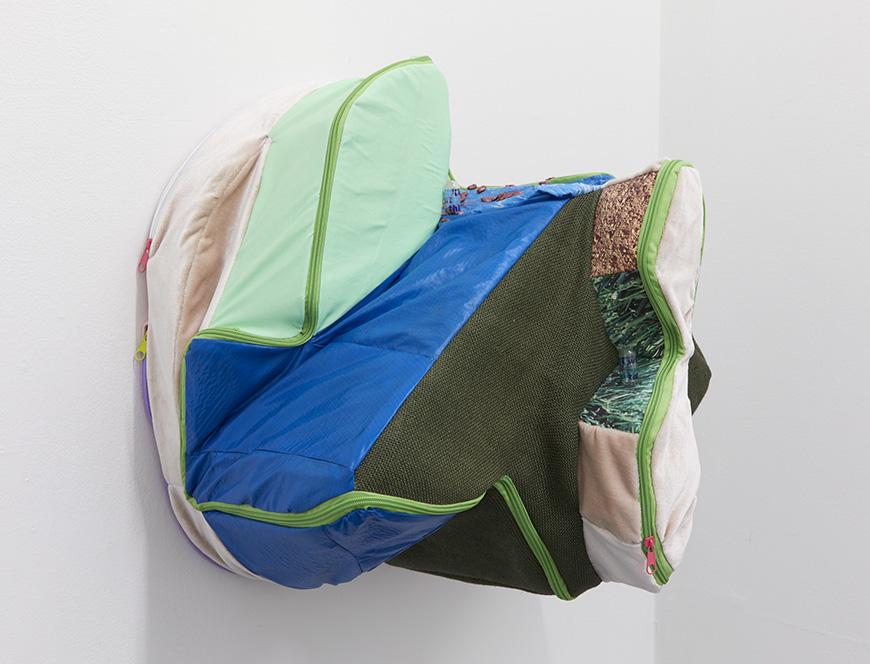 A three-dimensional fabric installation attached to a wall, made up of fabrics in many different colors, textures, and shapes, with a light green zipper running throughout.